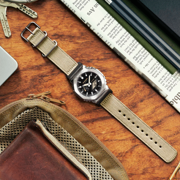 Reloj Casio G-Shock de acero con esfera negra, exterior metálico y correa de tela en color marrón claro.
